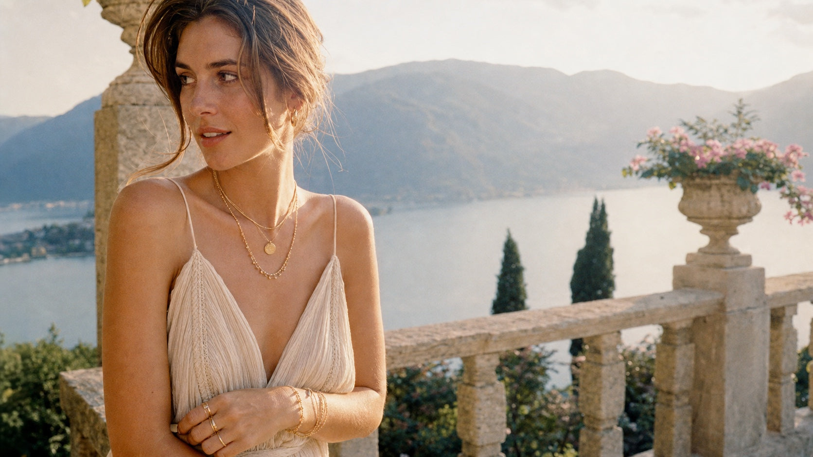 Model wearing layered Suri solid gold jewelry on a sunlit lakeside terrace