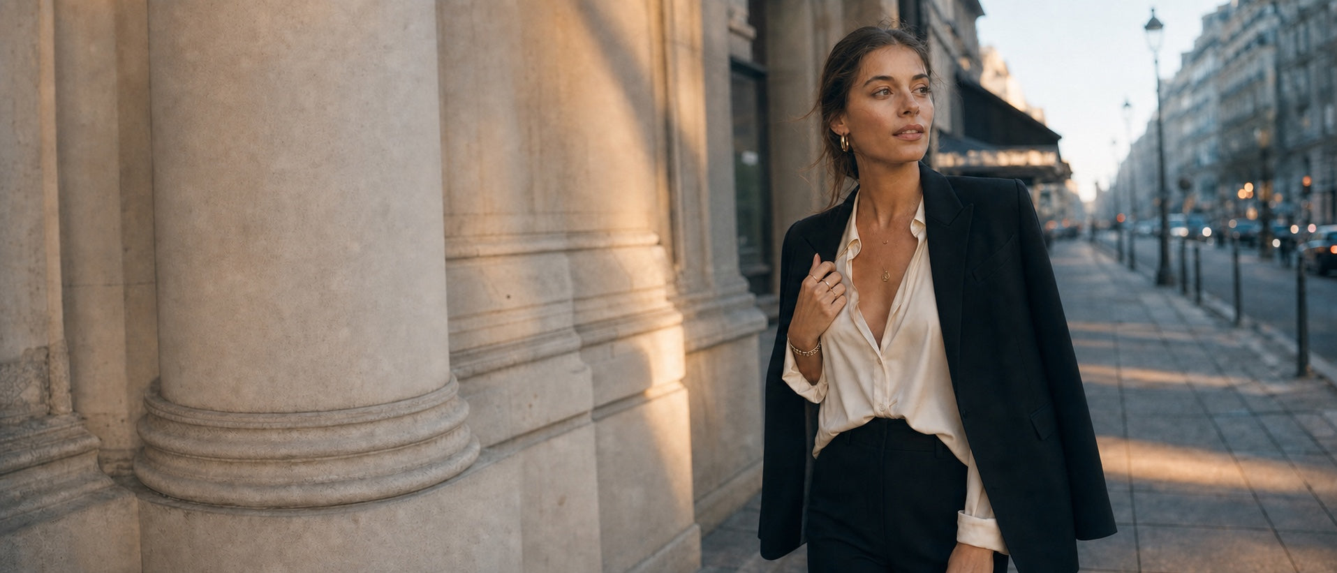Model wearing layered Suri solid gold jewelry and a tailored black outfit beside sunlit city columns