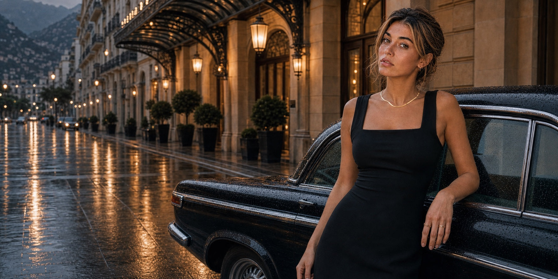 Model wearing Suri gold jewelry beside a vintage car outside a grand hotel after rain